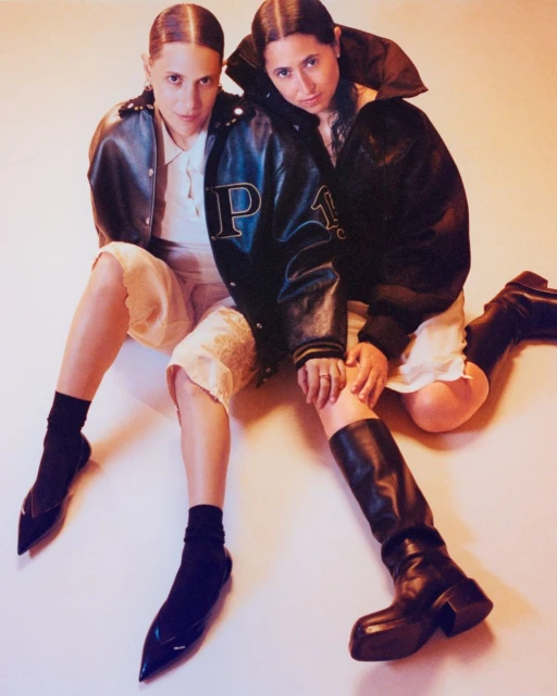 Two women in jackets and shorts sit side by side in a studio with warm lighting. One wears boots and the other pointed flats.