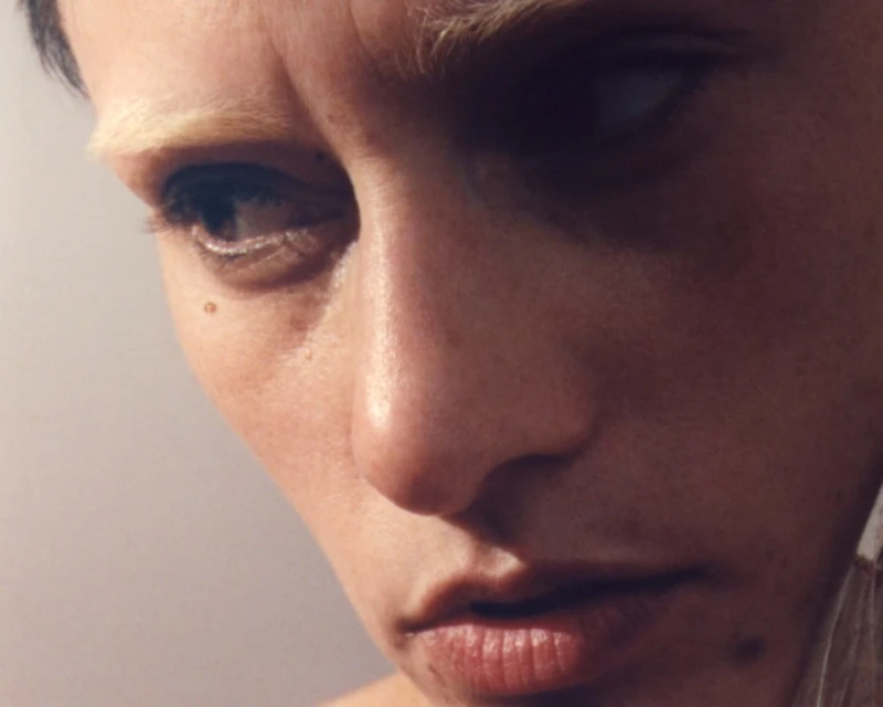 Close-up portrait of an adult female with shaved head and light eyebrows, looking away from the camera in a soft-lit, neutral setting.
