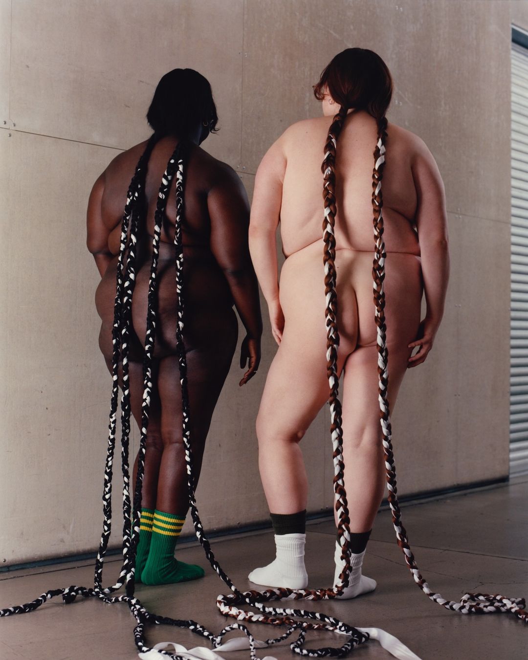 Two nude, plus-size women stand in front of a concrete wall. Their long braided extensions touch the floor, with green and white socks.