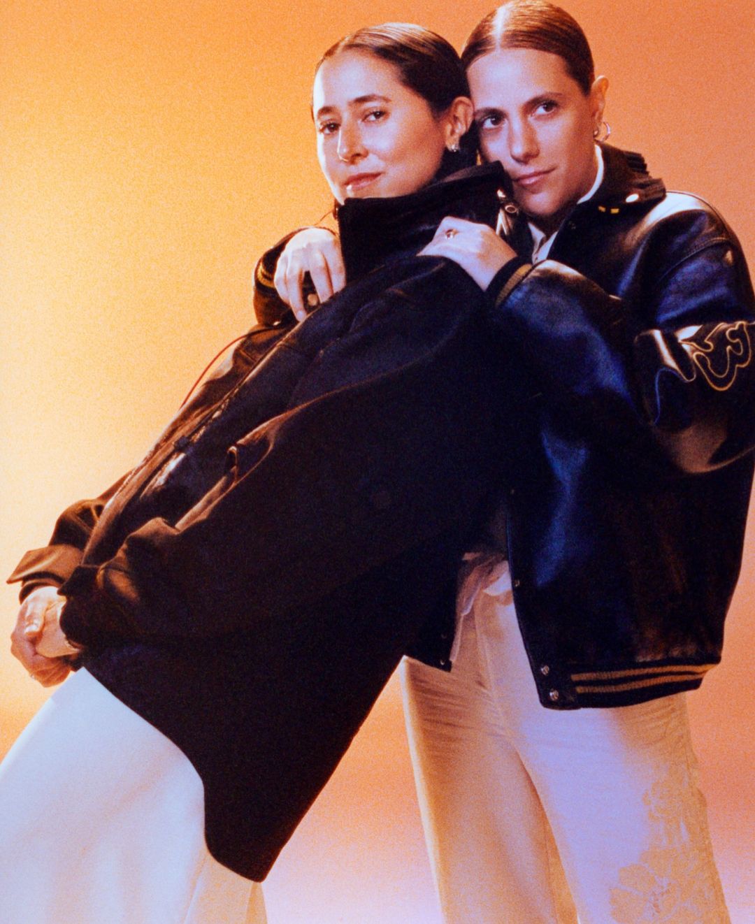 Two young women pose in a studio, wearing black leather jackets and cream-colored pants against a gradient orange background.