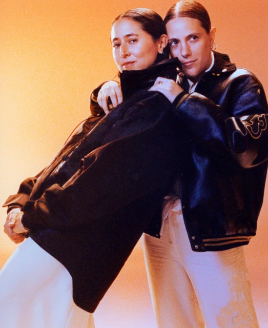 Two young women pose in a studio, wearing black leather jackets and cream-colored pants against a gradient orange background.