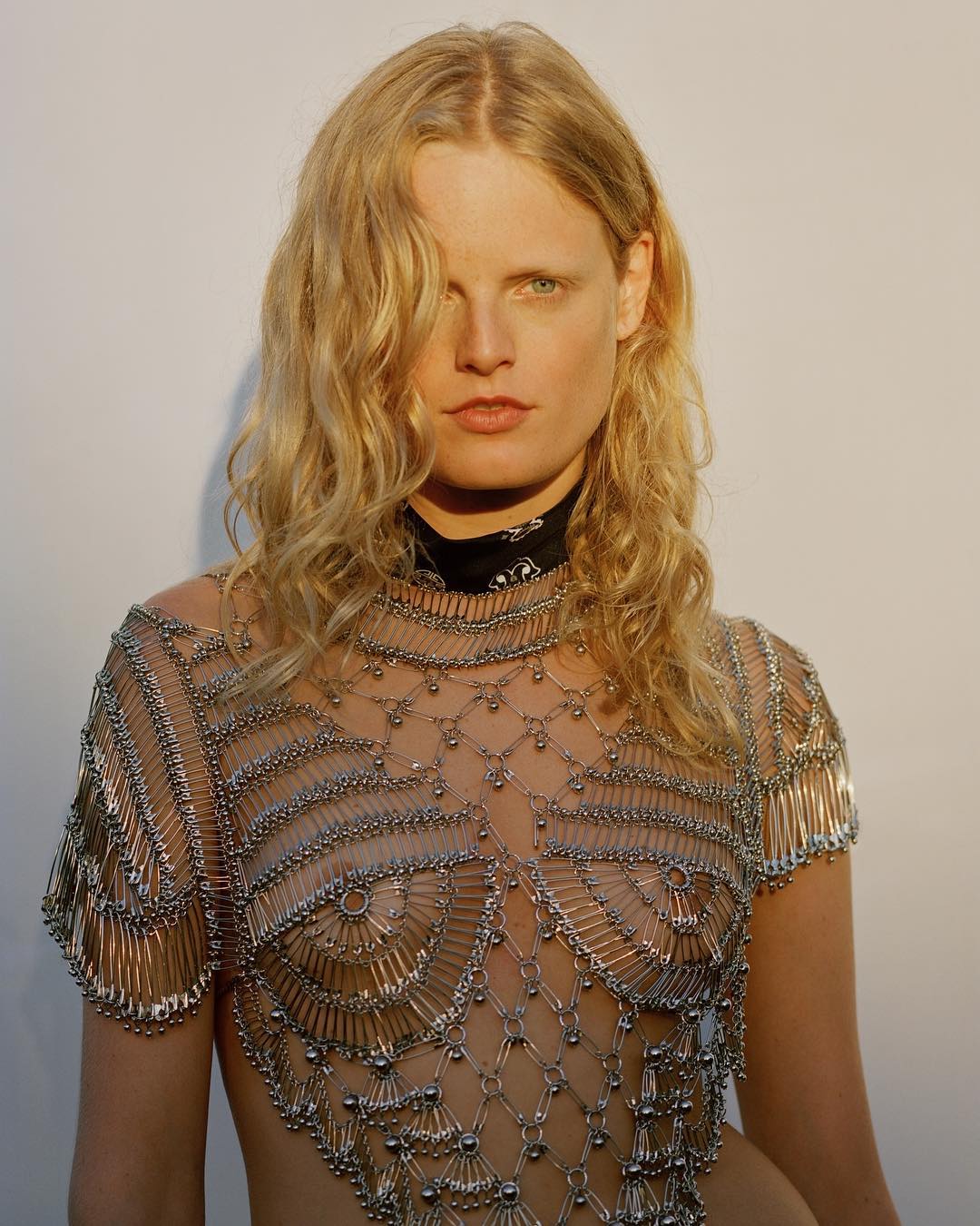Blonde model wears a top made of metal safety pins against a plain background in a fashion editorial shot with natural lighting.