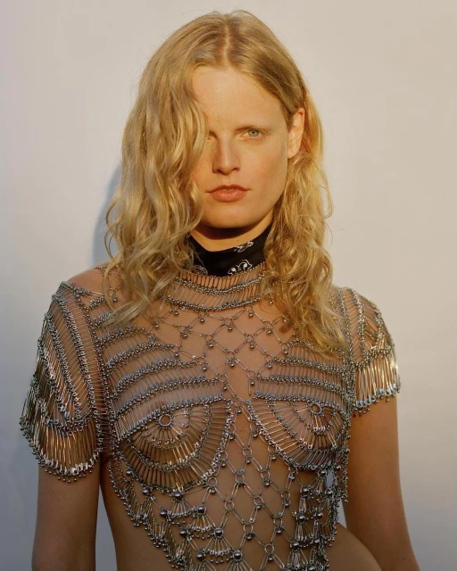 Blonde model wears a top made of metal safety pins against a plain background in a fashion editorial shot with natural lighting.