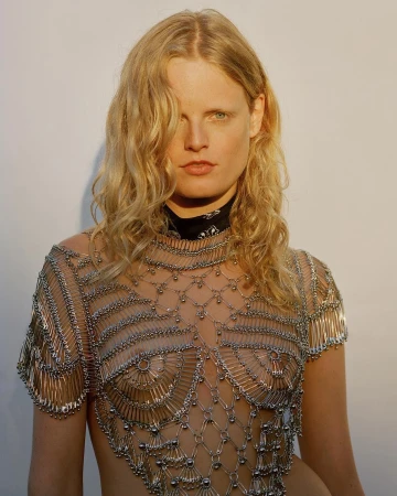 Blonde model wears a top made of metal safety pins against a plain background in a fashion editorial shot with natural lighting.
