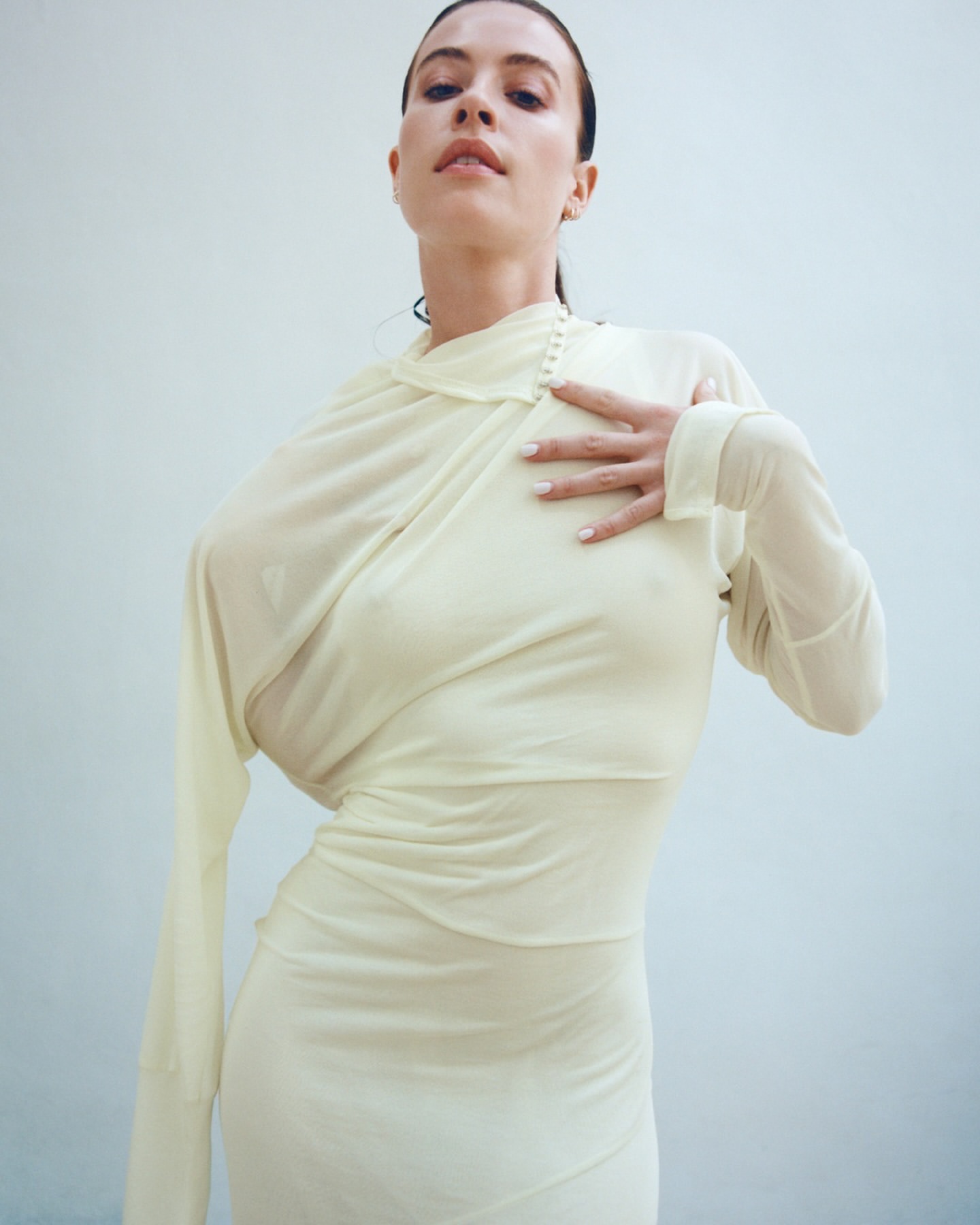 Elegant woman in sheer cream gown poses against a white backdrop. She has dark hair and her hand is on her chest, looking up.