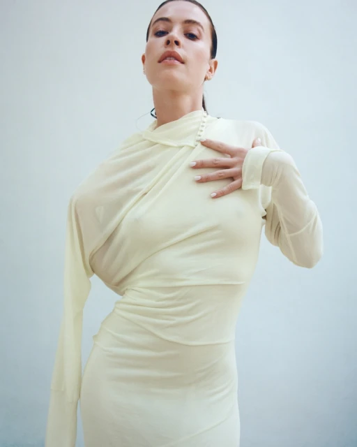 Elegant woman in sheer cream gown poses against a white backdrop. She has dark hair and her hand is on her chest, looking up.