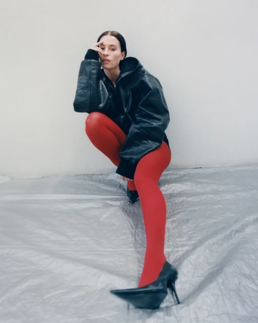 Woman squats in black jacket and red tights. She poses indoors in high heels on gray metallic material against a light wall.