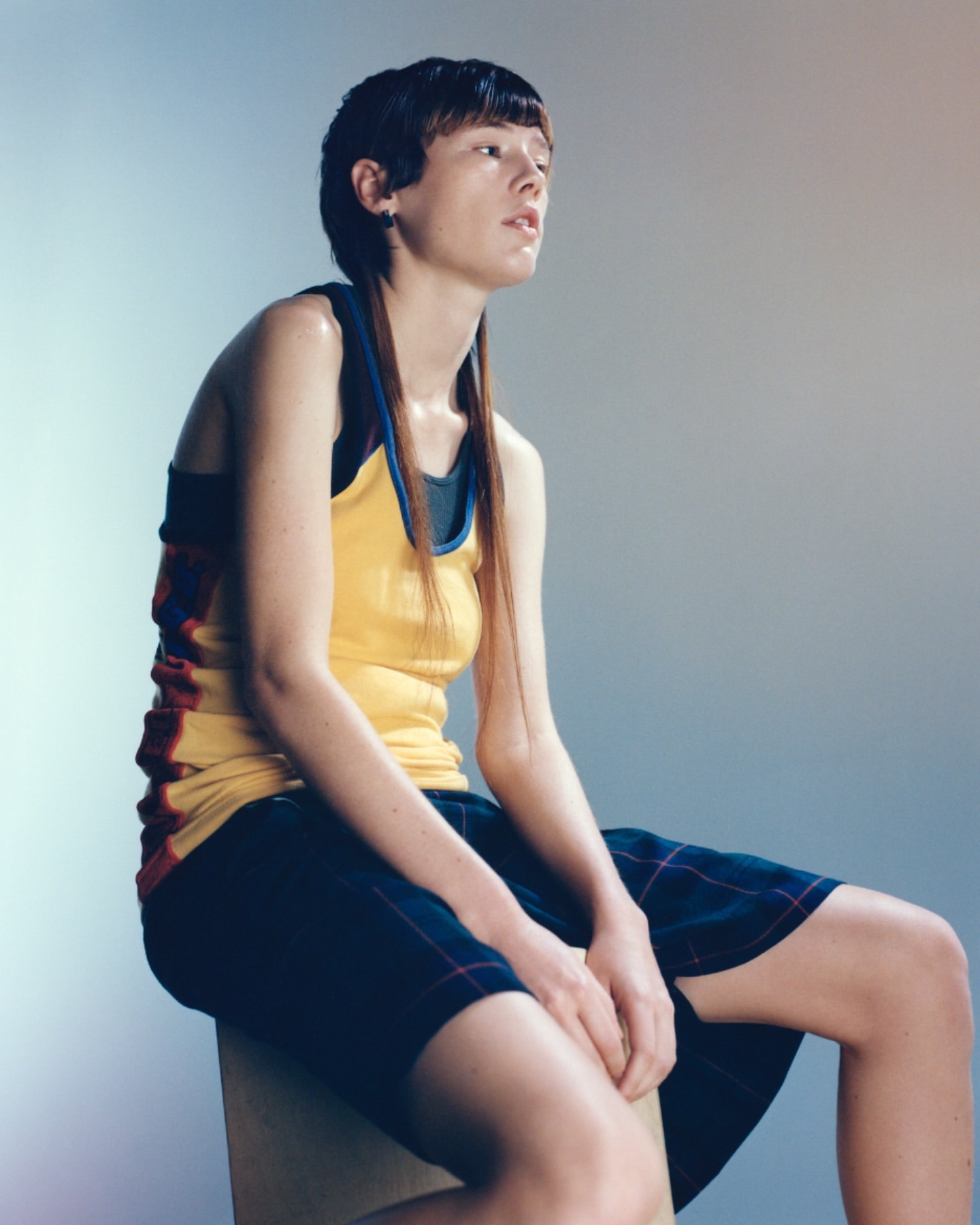 Adult female with a short haircut sits on a box, wearing a yellow top and plaid shorts, captured in a studio setting with soft, diffused light.