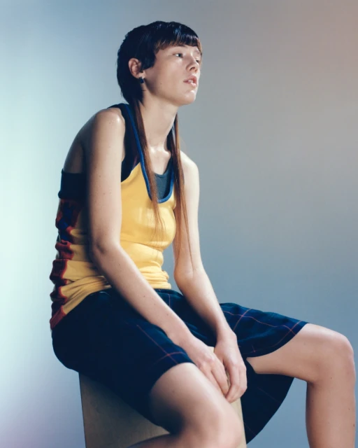 Adult female with a short haircut sits on a box, wearing a yellow top and plaid shorts, captured in a studio setting with soft, diffused light.