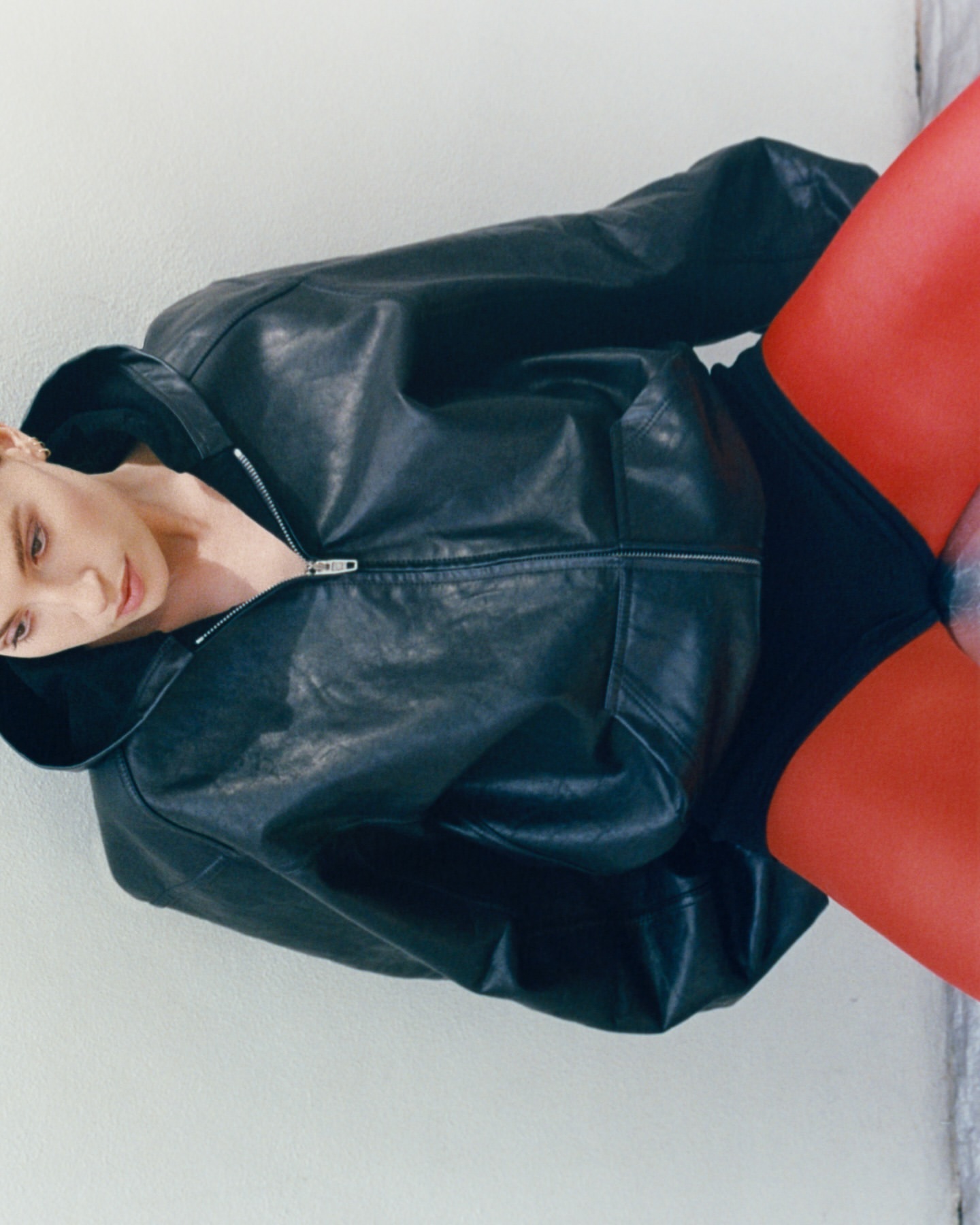 A woman with fair skin poses indoors wearing a black leather zip-up jacket, black undergarments, and bright red hosiery against a white wall.