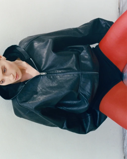 A woman with fair skin poses indoors wearing a black leather zip-up jacket, black undergarments, and bright red hosiery against a white wall.