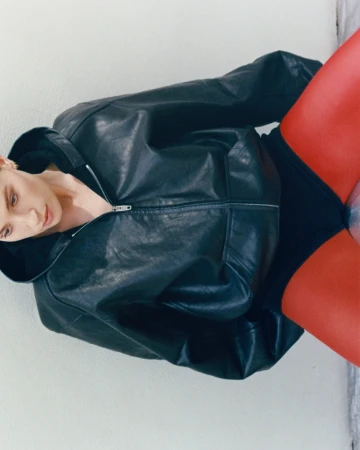 A woman with fair skin poses indoors wearing a black leather zip-up jacket, black undergarments, and bright red hosiery against a white wall.
