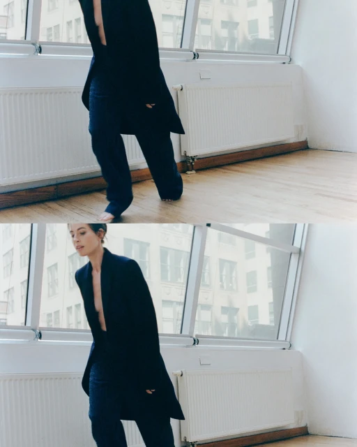 Adult woman in long coat and matching pants strikes a walking pose against a bright interior window with buildings visible outside.