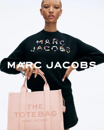 Model with shaved head wearing black sweater holding a pink tote bag against white background