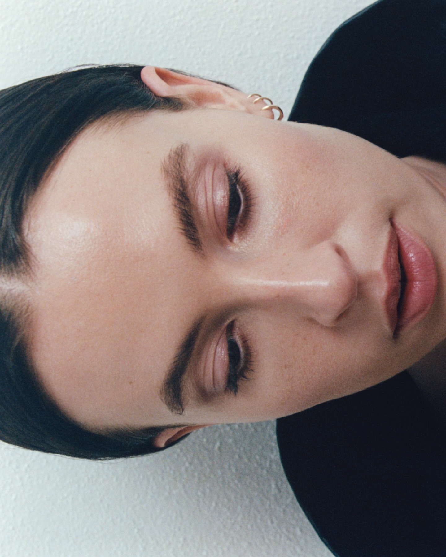 Close-up portrait of a woman with dark hair and gold hoop earrings against a white textured wall.