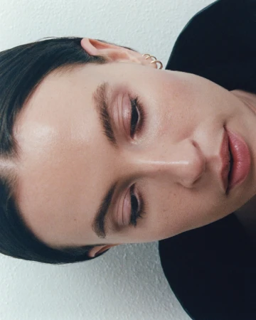 Close-up portrait of a woman with dark hair and gold hoop earrings against a white textured wall.