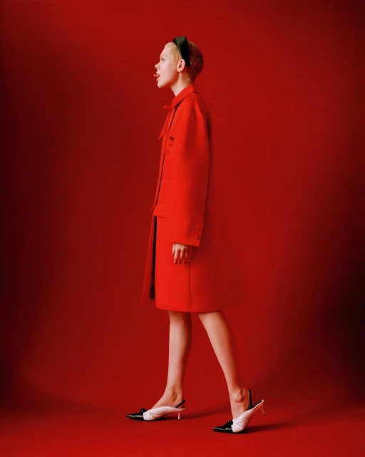 A woman in a red coat and black and white heeled shoes poses in profile against a matching red backdrop.