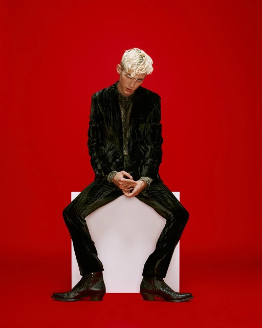 Male fashion model with blond hair wears a dark green velvet suit, sitting on a white cube against a solid red background in studio.