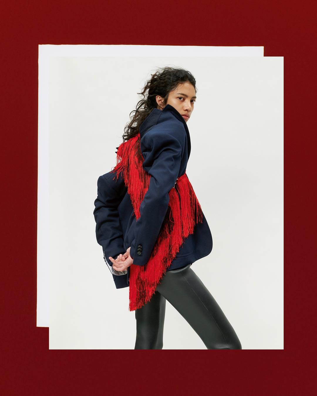 Fashion shot of a woman in a navy blazer with red fringe, black leggings, posing in a studio setting, framed by red and white.