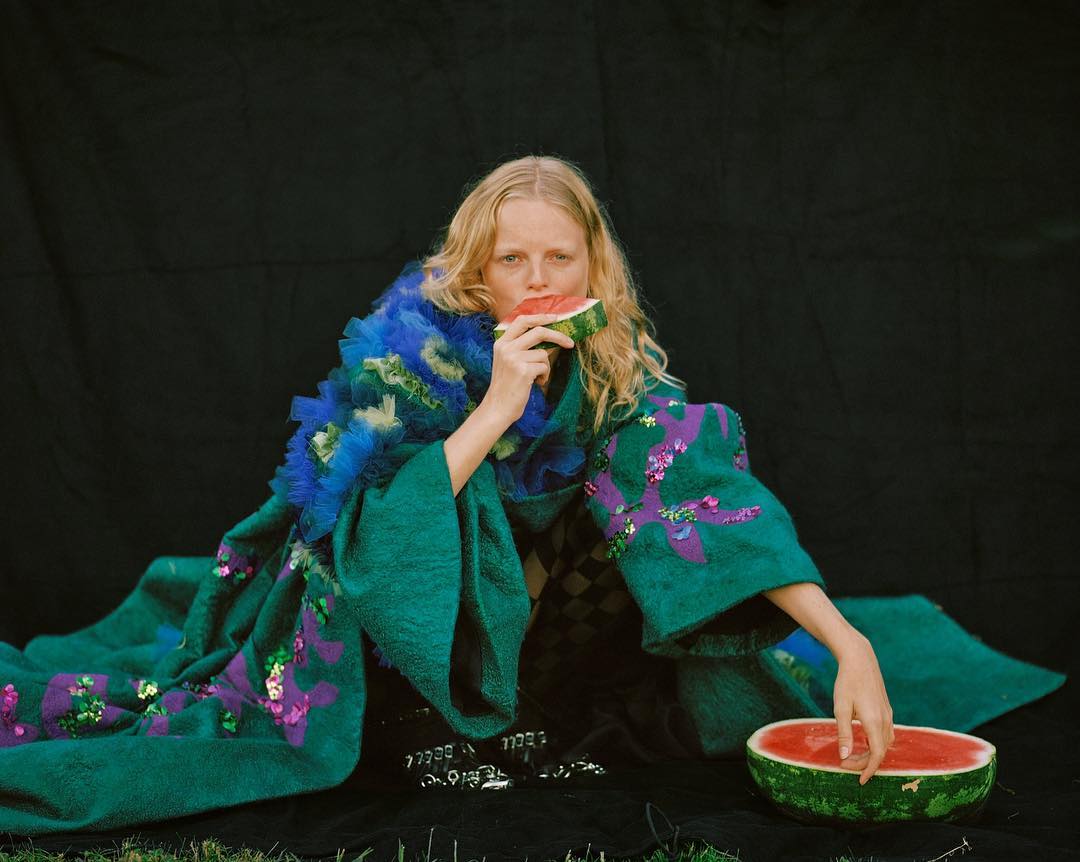 Fashion editorial shot: fair-skinned woman with watermelon, wearing emerald coat, posed on grass with a black background, eye-level view.