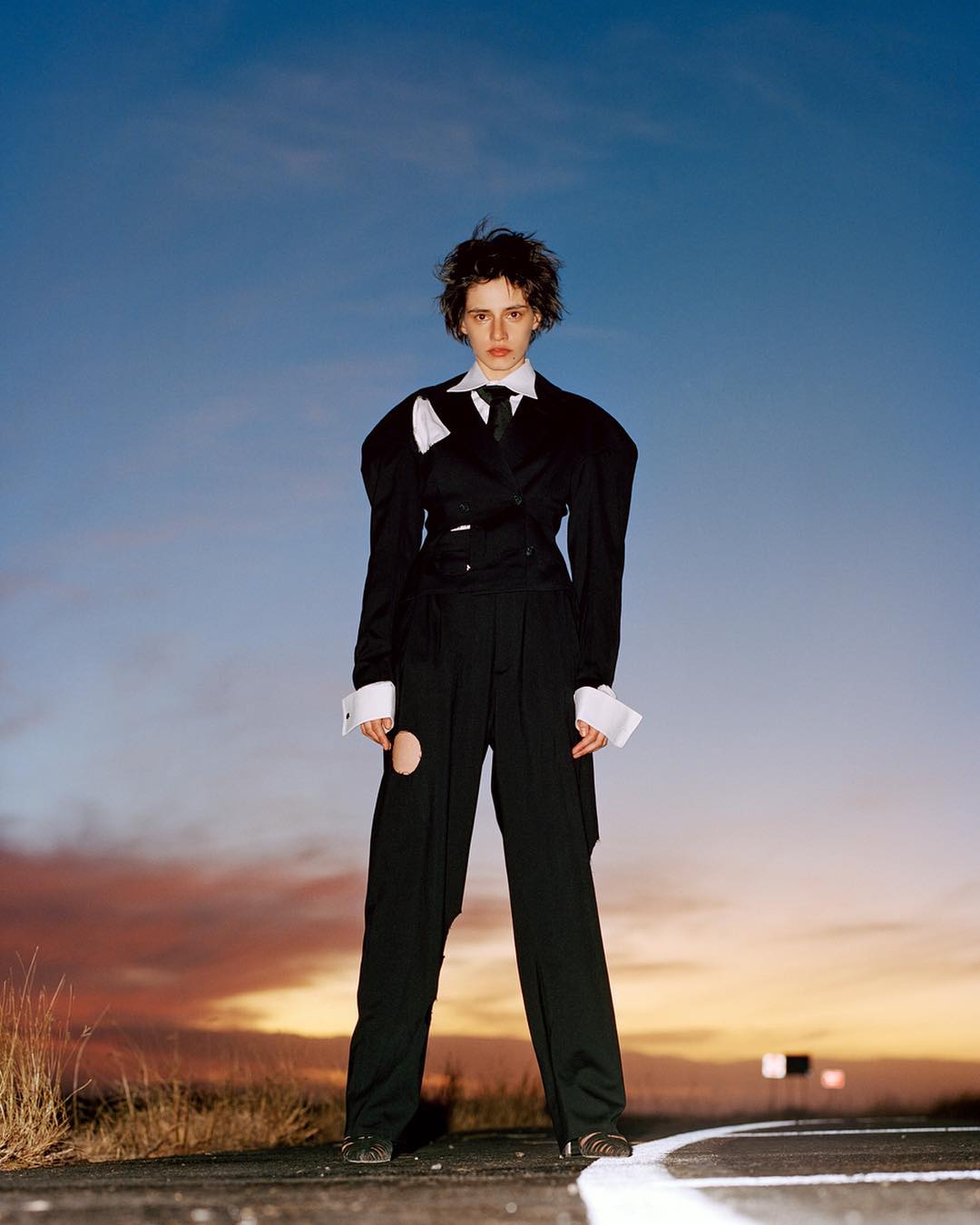 Stylish young adult woman in a black deconstructed suit posing outdoors at dusk against a blue and orange sky. Fashion editorial.