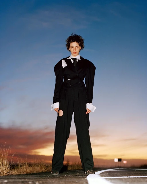 Stylish young adult woman in a black deconstructed suit posing outdoors at dusk against a blue and orange sky. Fashion editorial.