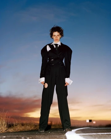 Stylish young adult woman in a black deconstructed suit posing outdoors at dusk against a blue and orange sky. Fashion editorial.