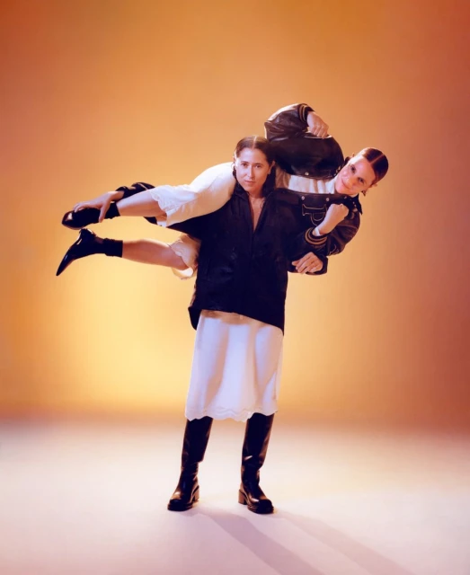 Two women in leather jackets and dresses; one is holding the other on her shoulder in a photo studio with a gradient orange background.