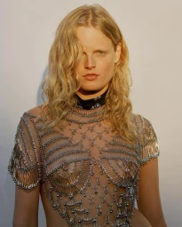 Model with blonde wavy hair wearing a metallic safety pin top against a plain wall photographed by Mat+kat
