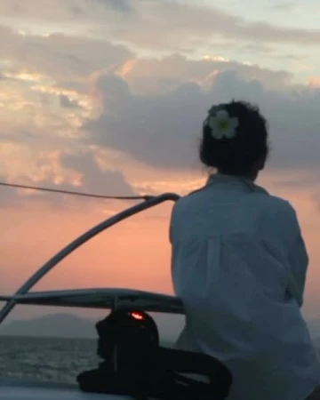 Woman in white shirt with flower in hair looking at sunset from boat deck by Maia Flore
