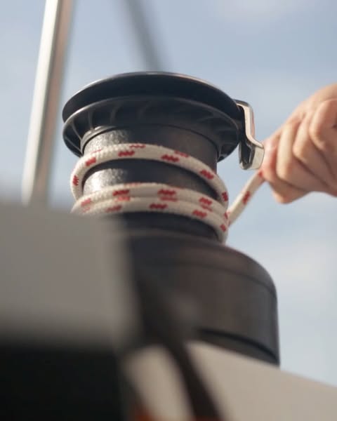 Close-up of a person's hand operating a boat winch with a white rope against a blue sky, evoking nautical activity.