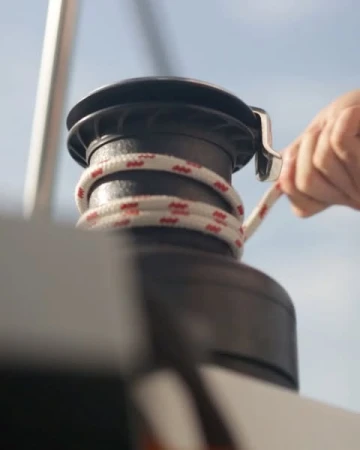 Close-up of a person's hand operating a boat winch with a white rope against a blue sky, evoking nautical activity.