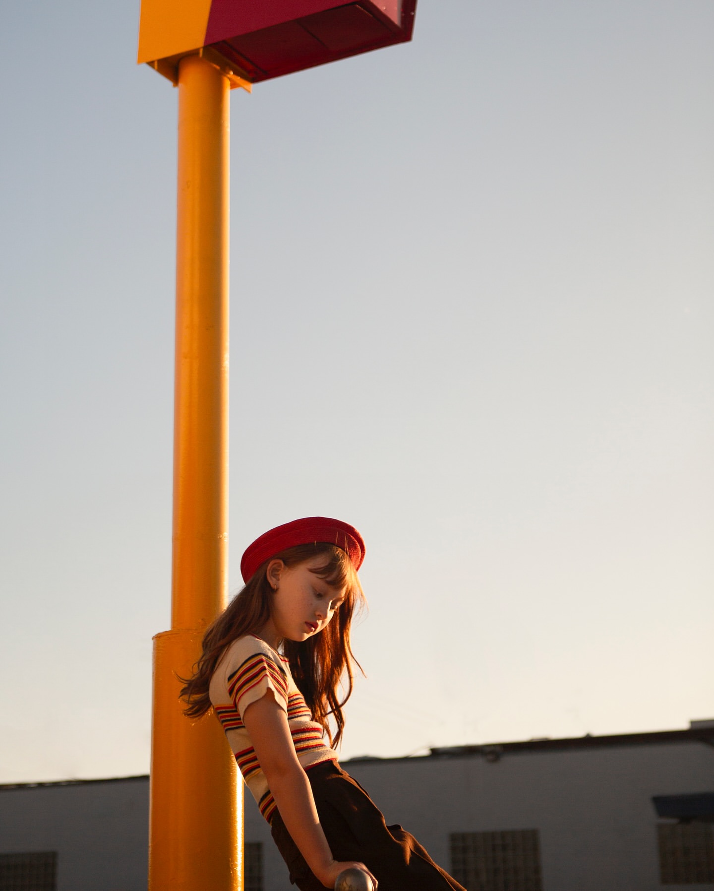 Young woman in a red beret leans against a yellow post in an urban setting, posing in a striped shirt and dark skirt under the open sky.