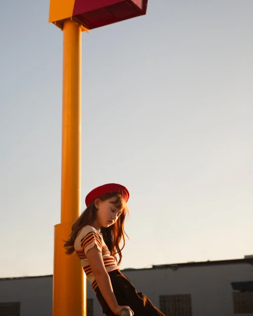 Young woman in a red beret leans against a yellow post in an urban setting, posing in a striped shirt and dark skirt under the open sky.