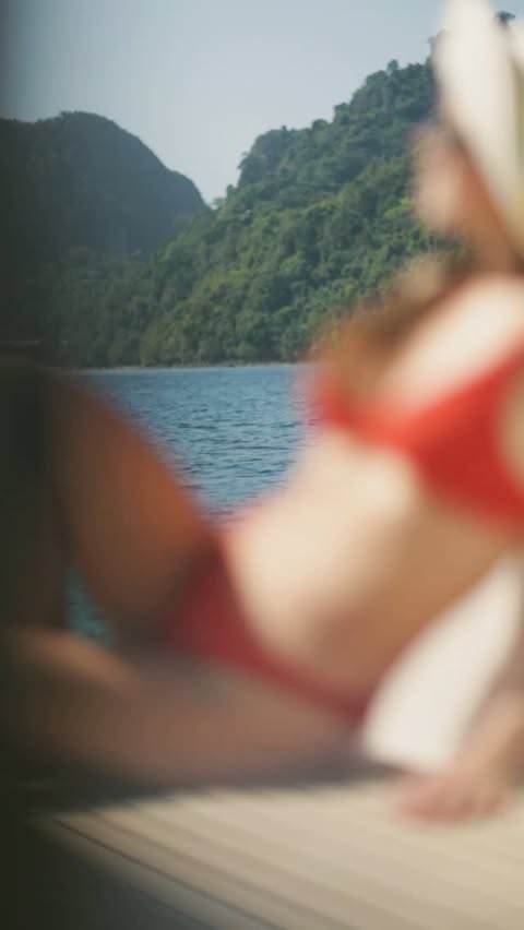Woman in red bikini lies on a wooden deck in front of the water and a mountainous, green, tropical island under a clear blue sky.