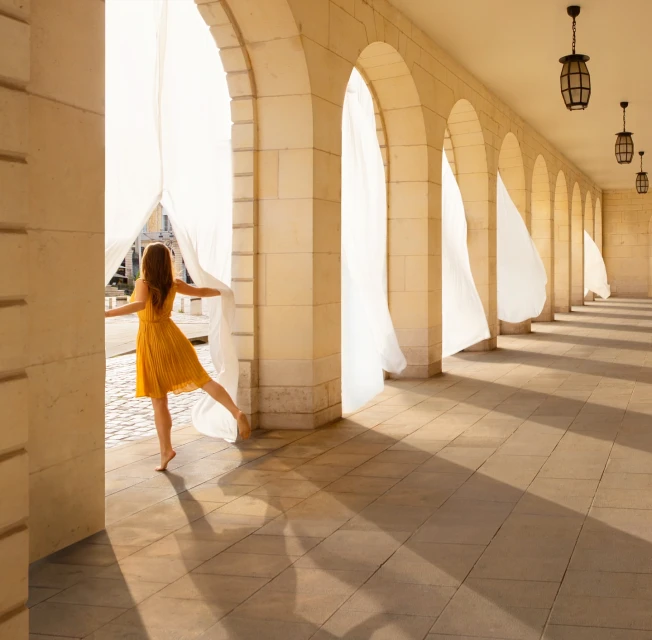 Woman in yellow dress stands in arched walkway with flowing white curtains in bright light, creating a sense of freedom and movement.