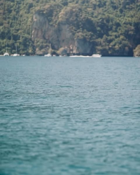 Calm turquoise ocean water meets a tree-covered rock formation in the distance under bright daylight. Boats are seen on the water.