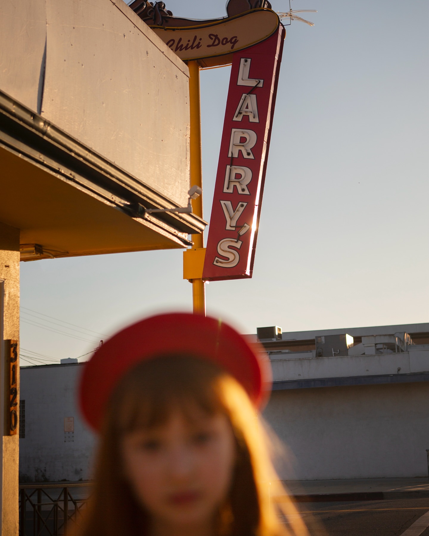 Young girl in red hat stands near vintage Larry's Chili Dog restaurant sign on a clear day with bright, warm light and muted colors.