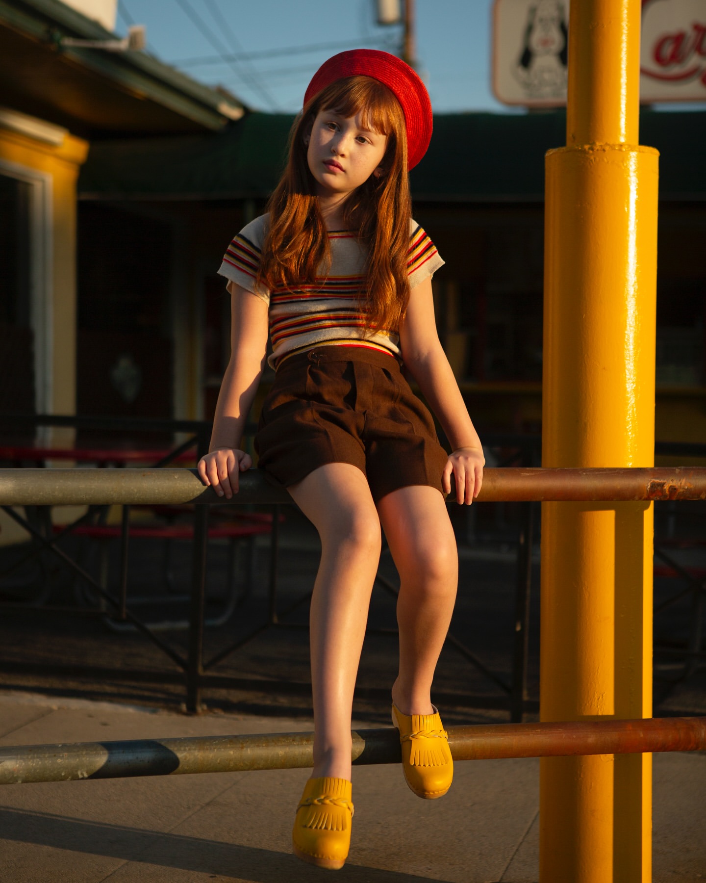 A young red-haired girl in a red beret sits on a railing wearing vintage-style brown shorts and a striped shirt in outdoor light.