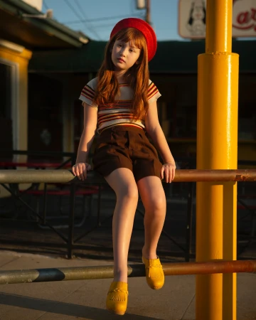 A young red-haired girl in a red beret sits on a railing wearing vintage-style brown shorts and a striped shirt in outdoor light.