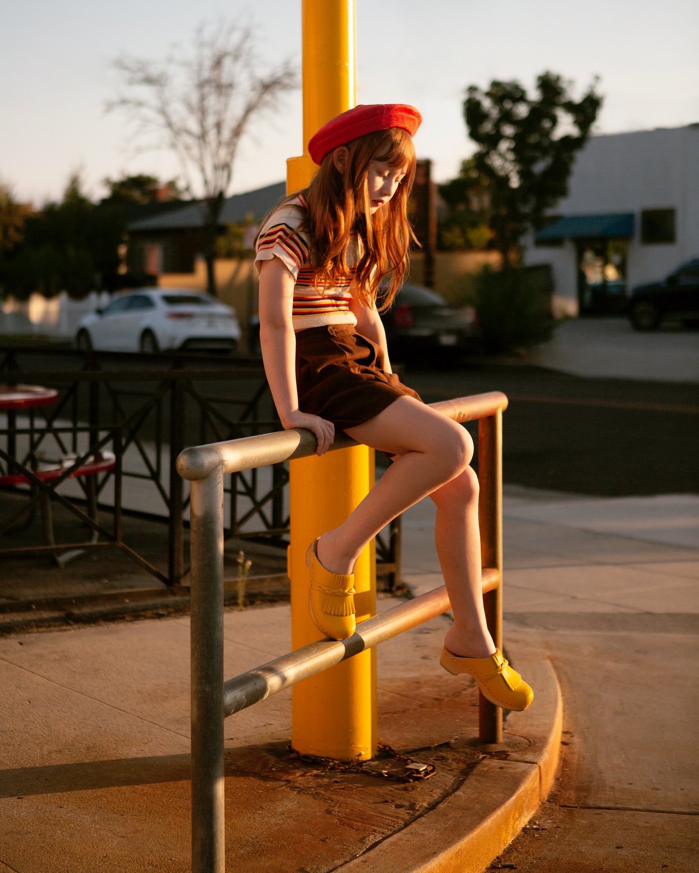 A young woman in a red beret and yellow shoes sits on a silver railing in a sunlit outdoor scene with cars and buildings in the background.