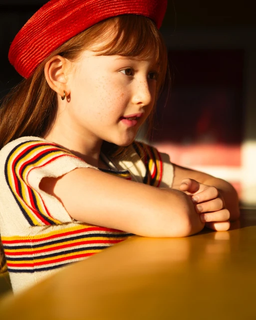 A young girl with red hair and freckles sits at a table in warm lighting, wearing a red beret and a striped short-sleeved shirt.