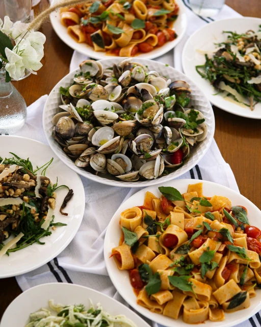 Overhead shot showcases an array of pasta dishes and shellfish, arranged on a wooden table and draped with a white linen cloth.