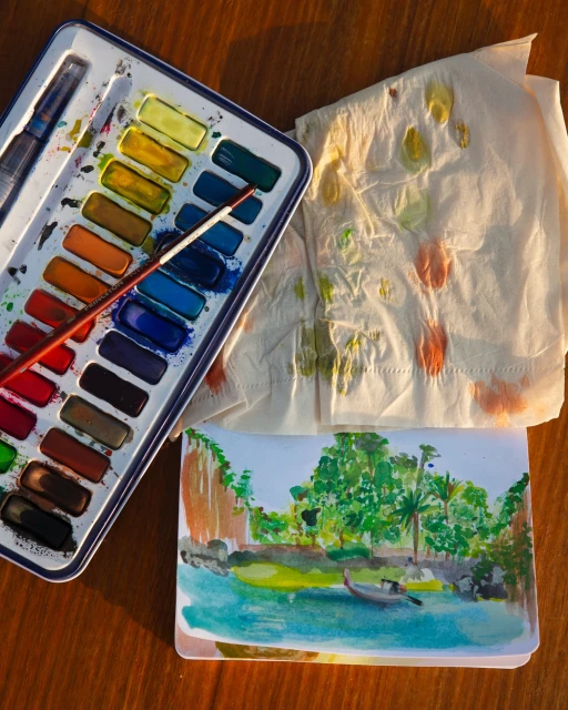 Watercolor painting on a wooden table with a travel scene, paint palette, paint brush, and paper towel with watercolor marks.