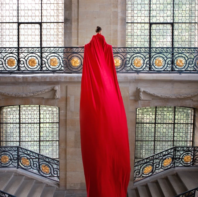 An adult female stands near a staircase wearing a long red gown in an indoor setting with large windows and decorative ironwork.