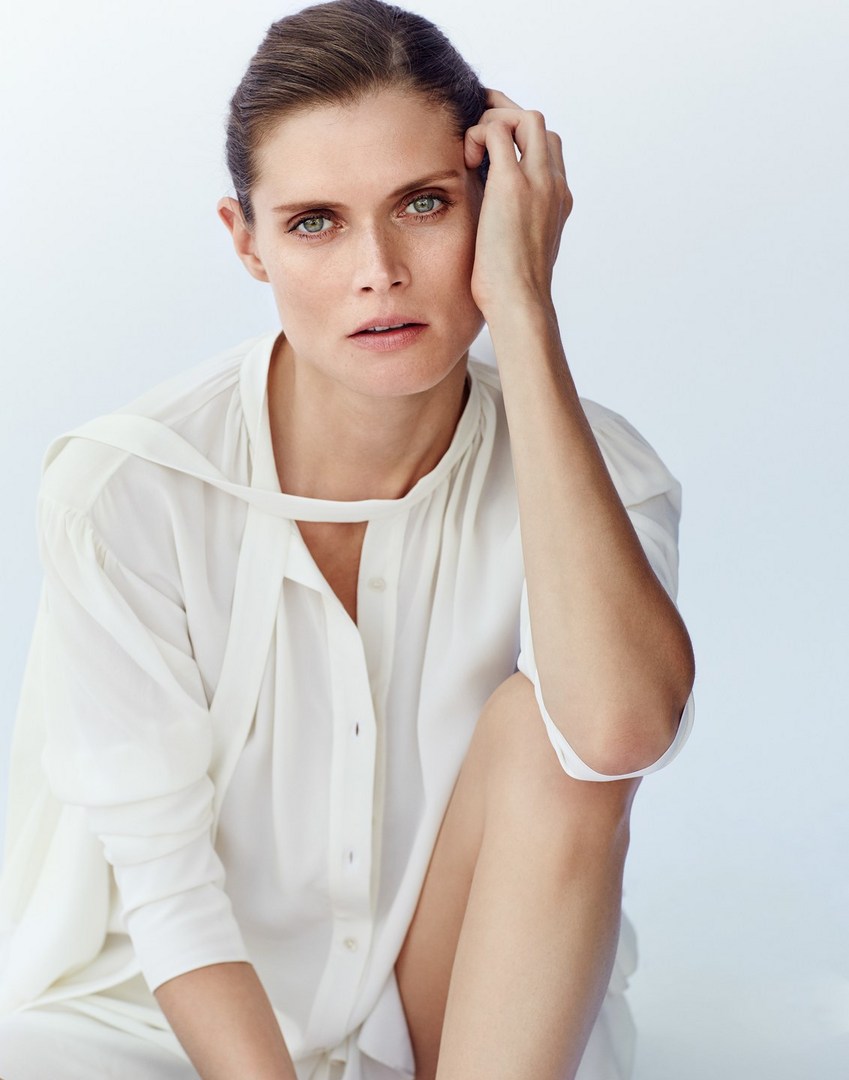 Close-up studio portrait of an adult woman with neutral expression, wearing a white button-down shirt, against a bright neutral backdrop.