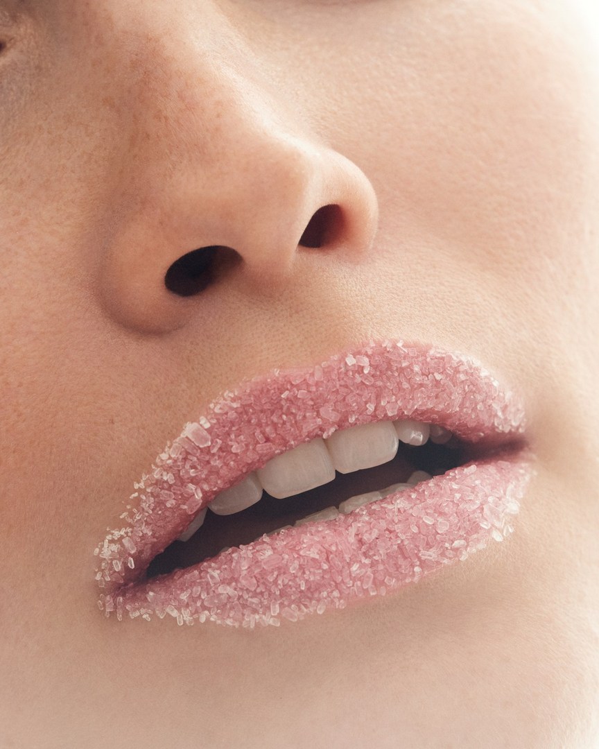 Close-up portrait of a woman's face featuring pink lips coated with sugar-like crystals, soft skin, and neutral background.