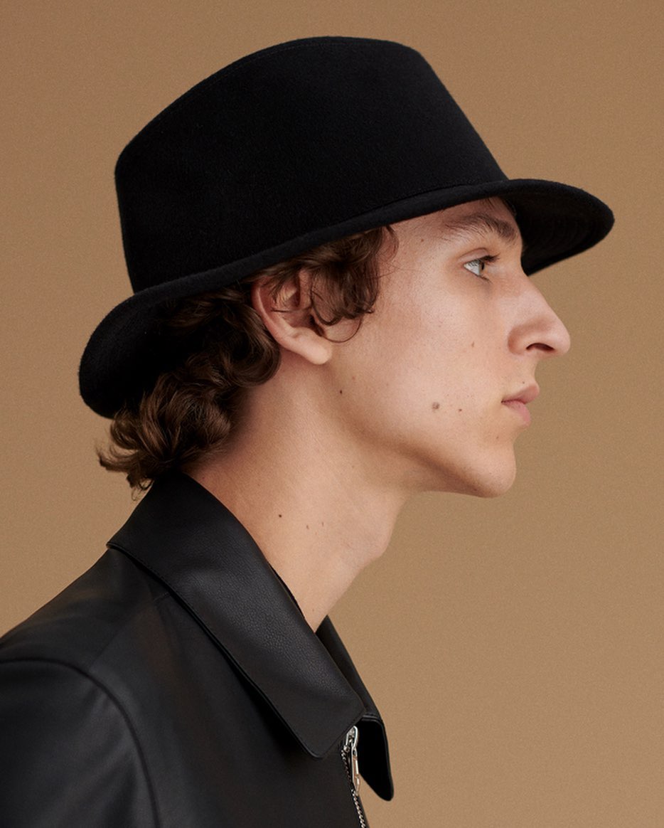 Profile portrait of an adult male with curly hair wearing a black hat and black jacket against a neutral beige background. 
