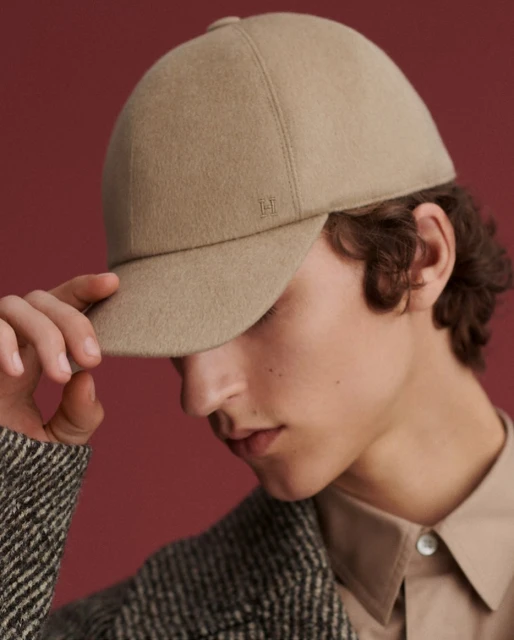 A young male model wears a tan wool baseball cap indoors, close-up shot, showcasing fashion and style against a solid reddish-brown background.