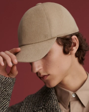 A young male model wears a tan wool baseball cap indoors, close-up shot, showcasing fashion and style against a solid reddish-brown background.